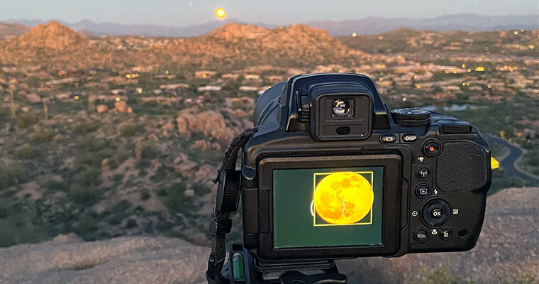 Camera on a tripod capturing a full moon over a rugged landscape at twilight.